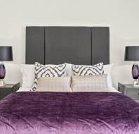 A modern bedroom with a dark grey headboard, white and patterned pillows, a purple bedspread, and two black nightstands with matching lamps on each side.