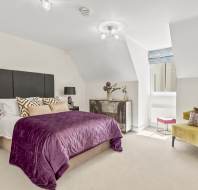 A modern bedroom with a double bed featuring a purple quilt, black headboard, two bedside tables with lamps, a dresser, an armchair, and a window with a Roman shade.