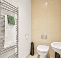 Modern bathroom with beige tiled wall, white toilet, wall-mounted sink, chrome towel rack holding two towels, and two decorative vases on the floor.
