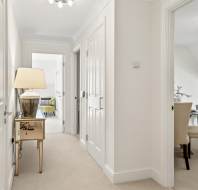 A bright hallway with beige carpet, a side table with a lamp, and doors leading to a bedroom and a dining area with cream chairs.