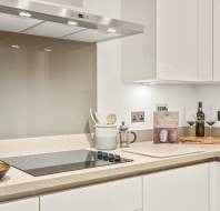 Modern kitchen with white cabinets, electric cooktop, utensils, a cookbook, wine bottle, two wine glasses, potted flowers, and a gold bowl on a beige countertop.