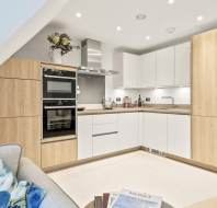 Modern L-shaped kitchen with wood and white cabinets, built-in appliances, a sink, and countertop under bright ceiling lights; part of a sofa and coffee table in foreground.