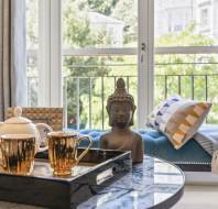 A living room with a marble coffee table featuring a tray with two gold mugs and a teapot, a Buddha statue, a bench with cushions, and large windows showing greenery outside.