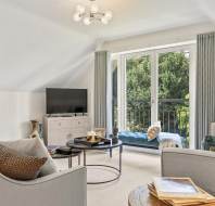 Bright living room with a TV, two sofas, a coffee table, books, and a window seat overlooking greenery through large glass doors.