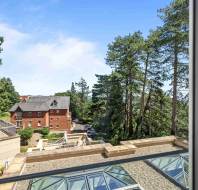 View from a window showing a courtyard with gravel and glass panels, a red brick building, parked cars, and tall trees under a blue sky.