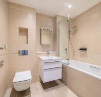 Modern bathroom with beige tiles, wall-mounted toilet, heated towel rail, white vanity with sink, mirror, and a bathtub with glass shower screen and overhead shower.