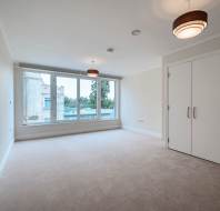 Empty, carpeted room with large window, two ceiling lights, double doors, and neutral-colored walls. View of buildings and trees outside.