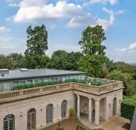 View of a neoclassical building with arched windows and a rooftop garden, surrounded by trees under a partly cloudy sky.