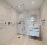 Modern, accessible bathroom with walk-in shower, wall-mounted grab bars, handheld showerhead, glass partition, and a white vanity with mirror. White tiled walls and beige tiled floor.