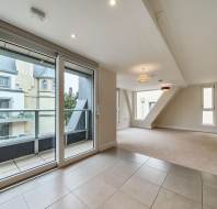Modern, unfurnished apartment interior with large windows, balcony access, and a mix of tiled and carpeted floors. Neutral walls and ceiling lights are visible.
