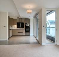 Modern apartment interior with open-plan kitchen, tiled and carpeted flooring, large windows, and a glass door leading to a balcony.