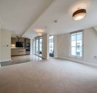 Unfurnished modern apartment with open-plan living area, beige carpet, large windows, and a kitchen with built-in appliances and tile flooring.