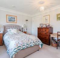 A bedroom with a bed, patterned quilt, wooden nightstands, a wooden dresser with photos, a desk with a chair, framed artwork on the walls, and a ceiling light fixture.