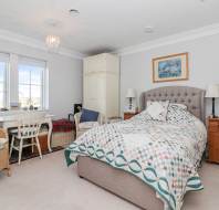 A neatly arranged bedroom with a bed, wicker chair, desk by the window, wardrobe, side tables, and a standing lamp, decorated in neutral tones with a patterned quilt on the bed.