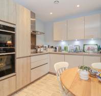 Modern kitchen with light wood cabinets, built-in double ovens, countertop appliances, dish rack, and a round wooden table set with fruit and a plant.