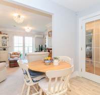 A cozy dining area with a round wooden table and four chairs is adjacent to a bright living room featuring sofas, cabinets, and large windows letting in natural light.