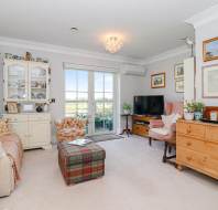 A cozy living room with beige carpet, armchairs, a TV on a stand, wooden furniture, framed pictures on the walls, and glass doors leading to a balcony with an outdoor view.