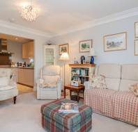 A cozy living room with two armchairs, a sofa, a plaid ottoman, bookshelf, framed pictures on the wall, and an open view into a modern kitchen.