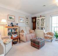 Bright living room with armchairs, a plaid ottoman, bookshelf, display cabinet, framed art on walls, and a large window with a view outside.