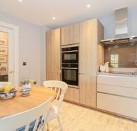Modern kitchen with light wood cabinets, built-in ovens, a round dining table with four chairs, and countertop appliances under a stainless steel range hood.