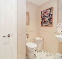 Modern bathroom with beige tiles featuring a toilet, wicker laundry basket, white sink, various toiletries, and a blurred wall picture above the toilet.