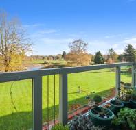 View from a balcony with potted plants, overlooking a green lawn, trees, and a pond under a clear blue sky.