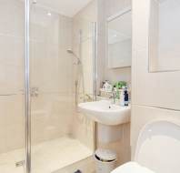 Modern bathroom with beige tiles, a glass shower enclosure, wall-mounted sink, various toiletries on the sink, a mirror cabinet, and a white toilet in the foreground.