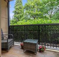 A small balcony with wicker chairs and a table, potted flowers, wooden flooring, and a decorative metal railing, overlooking green trees.