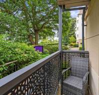 Small balcony with decorative metal railing, a wicker chair, and views of greenery and trees beside an apartment building.