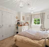 A neatly arranged bedroom with a double bed, light-colored furniture, closed closet doors, a window with curtains, and decorative items on shelves and dressers.