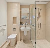 Modern bathroom with beige tiles, wall-mounted toilet and sink, walk-in glass shower, and towel radiator on the left wall.