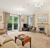 A cozy living room with beige sofas, a coffee table, a TV, a fireplace with ornaments, large windows with curtains, and various framed pictures on the walls.