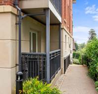 Ground-level apartment balcony with metal railing beside a paved walkway, surrounded by greenery and bordered by a brick and stone building.