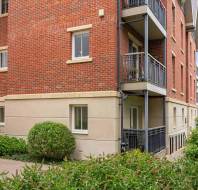 A red brick apartment building with balconies and trimmed bushes in front, seen on a clear day.