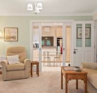 A cozy living room with beige armchairs, a wooden table, and a lamp, leading into a kitchen through glass double doors.