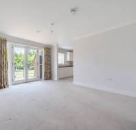 Unfurnished living room with beige carpet, floral curtains on glass doors leading outside, and an open view into a white kitchen area.
