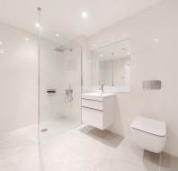 Modern, minimalistic bathroom with white tiles, a walk-in glass shower, wall-mounted sink with mirror, and a wall-mounted toilet. The space is well-lit and uncluttered.