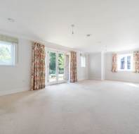 Spacious, empty room with beige carpet, white walls, windows on three sides, floral curtains, and glass doors opening to a garden.