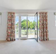 A bright room with floral curtains and two small windows, featuring open glass doors leading to a patio and a view of trees outside.