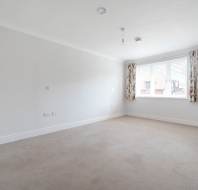 Empty, carpeted room with white walls, a window with floral curtains, and several electrical outlets visible on the left wall.