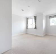 Empty, unfurnished white room with beige carpet, two windows with patterned curtains, and a single exposed ceiling light bulb.