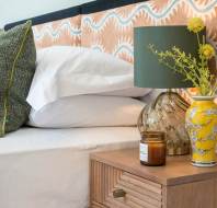 A bedside table with a green lamp, a yellow vase with flowers, and a candle next to a bed with white sheets and green pillows. Geometric headboard in background.