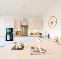 Modern kitchen with white cabinets, built-in appliances, a large island countertop, decorative items, and an open cookbook; dining area is visible in the background.