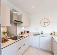 Modern kitchen with light cabinets, built-in oven, electric cooktop, sink, and minimalist decor including a wall clock and vase with dried plants on the countertop.