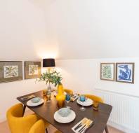 A dining area with a dark wooden table set for four, yellow chairs, yellow vases, and framed botanical art on white walls under sloped ceiling.