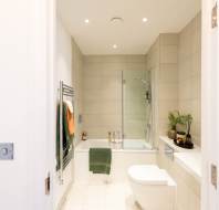 Modern bathroom with beige tiles, a bathtub and shower, a glass screen, a wall-mounted toilet, towel rack with towels, and a shelf with potted plants and decor items.