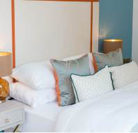Modern bedroom with a bed made up in white linens, decorative pillows, and two bedside tables holding matching lamps with beige shades and amber glass bases.