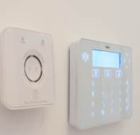 A close-up view of a wall-mounted security system control panel and a thermostat on a white wall.