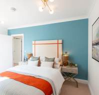 A modern bedroom with a blue accent wall, double bed with white and orange bedding, two bedside tables with lamps, and framed artwork on the walls.