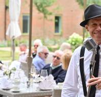 A man in a hat and suspenders stands with a vintage microphone, smiling, while people sit at outdoor tables in the background.